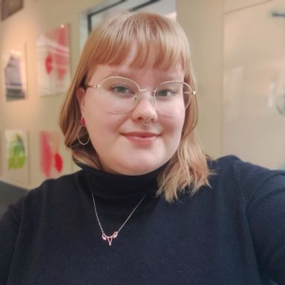 Kuvassa on Selmi Holopainen laajassa kasvokuvassa. Hänellä on musta asu, silmälasit ja vaaleat hartiapituiset hiukset otsatukalla. Taustalla näkyy taidenäyttelyn teoksia.