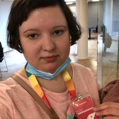 Selfiessä tummahiuksinen ja vaaleanpunaiseen paitaan pukeutunut henkilö nostaa esiin sateenkaarikaulanauhaa, jossa on vasemmistoliiton kokousedustaja-nimikortti.