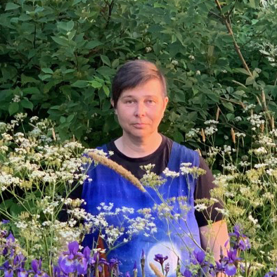 Ulla Kommonen seisoo niityllä. Etualalla näkyy sinisiä kukkia, taempana valkoisia ja taustalla pensaikkoa. Ulla katsoo suoraan kameraan, ja hänellä on lyhyet hiukset ja sininen asu.