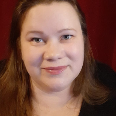 kasvukuva Outi Einistöstä, jolla on ruskeat pitkät hiukset ja musta paita. Taustalla näkyy punertavaa seinää.