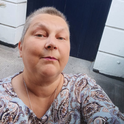 Selfie Eevi Kiimamaasta. Hänellä on lyhyet hiukset, sinikuviollinen paita ja kaulakoru. Taustalla on ovi ja valkoistä seinää.