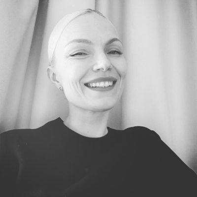 Mustaan paitaan pukeutunut Minja Koskela katsoo nauraen kameraan mustavalkokuvassa.