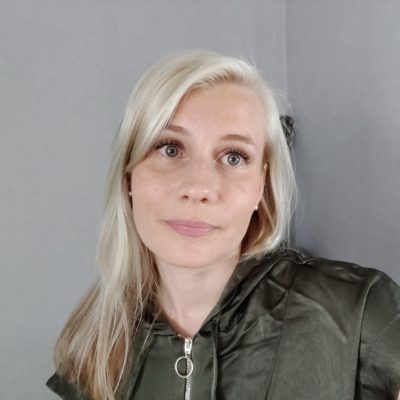 Jenny Simojoella on pitkät vaaleat hiukset ja hän on pukeutunut tummaan takkiin. Taustalla näkyy harmaata seinää.