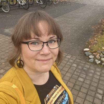 Selfiekuvasssa Jonna Parviainen seisoo kadulla. Takana näkyy pyöriä telineissään. Jonna on pukeutunut keltaiseen takkiin, hänellä on silmälasit, korvikset, printti-t-paita ja laukku olalla.