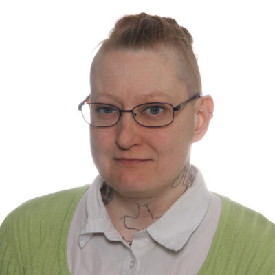 Silmälasipäinen, lyhythiuksinen, tatuoitu ja valkoiseen lkauluspaitaan ja vihreään neuleeseen pukeutunut Nina Hansen katsoo suoraan kameraan. Valokuvan tausta on rajattu pois.