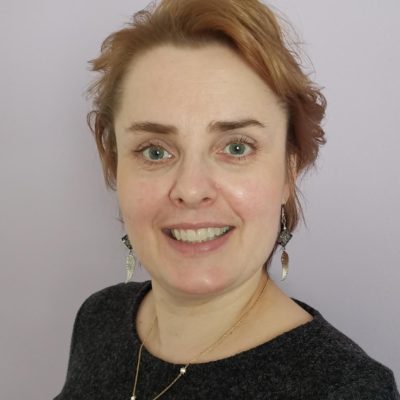 Kuvassa Petriina Punna katsoo kameraan hymyillen. Hänellä on lyhyet punertavat hiukset, kaulakoru ja korvakorut ja harmaa paita.